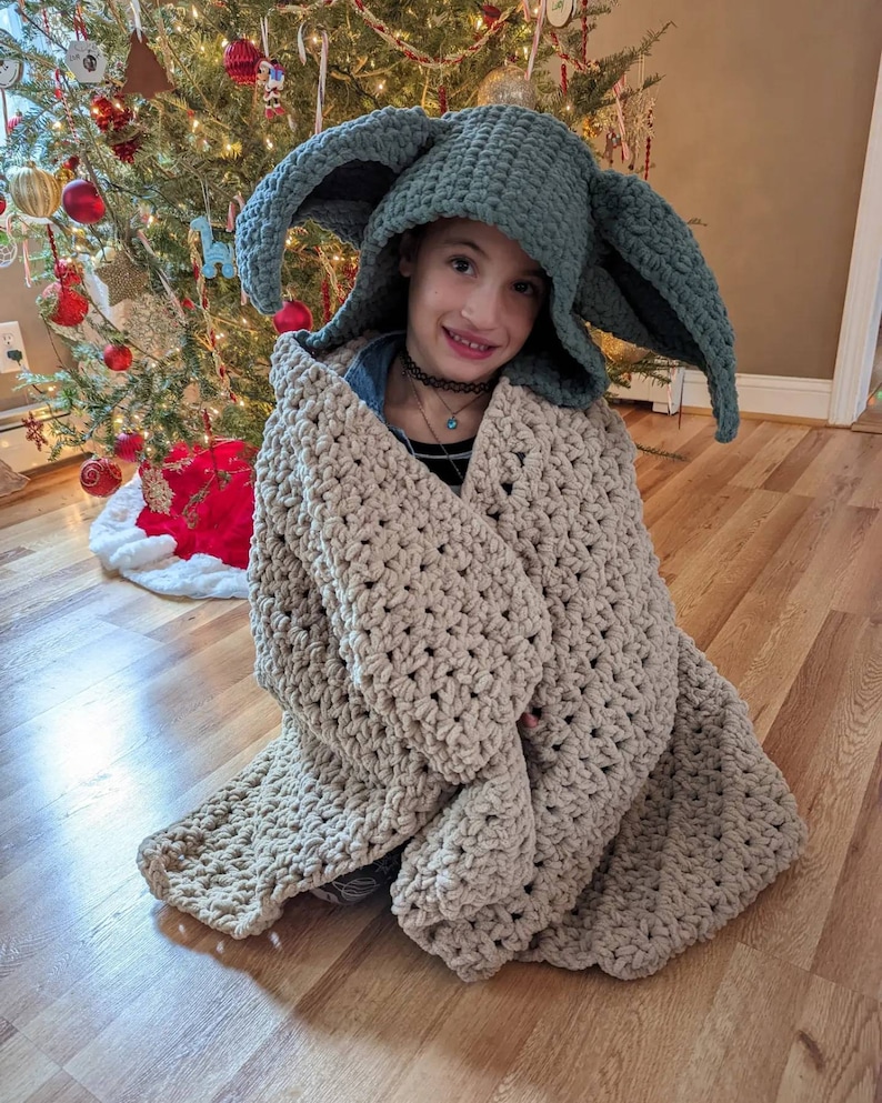 Puede incluir: Un ni&ntilde;o lleva un gorro verde tejido con grandes orejas puntiagudas y est&aacute; envuelto en una manta beige tejida. El gorro se asemeja a un personaje de una popular franquicia de ciencia ficci&oacute;n. La manta es gruesa y texturizada. Un &aacute;rbol de Navidad decorado est&aacute; en el fondo.