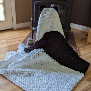 Puede incluir: Una manta con capucha de oso panda hecha a mano, blanca y marr&oacute;n. La manta est&aacute; hecha de un material suave y afelpado. La capucha presenta orejas de panda. La manta est&aacute; doblada sobre un suelo de madera.