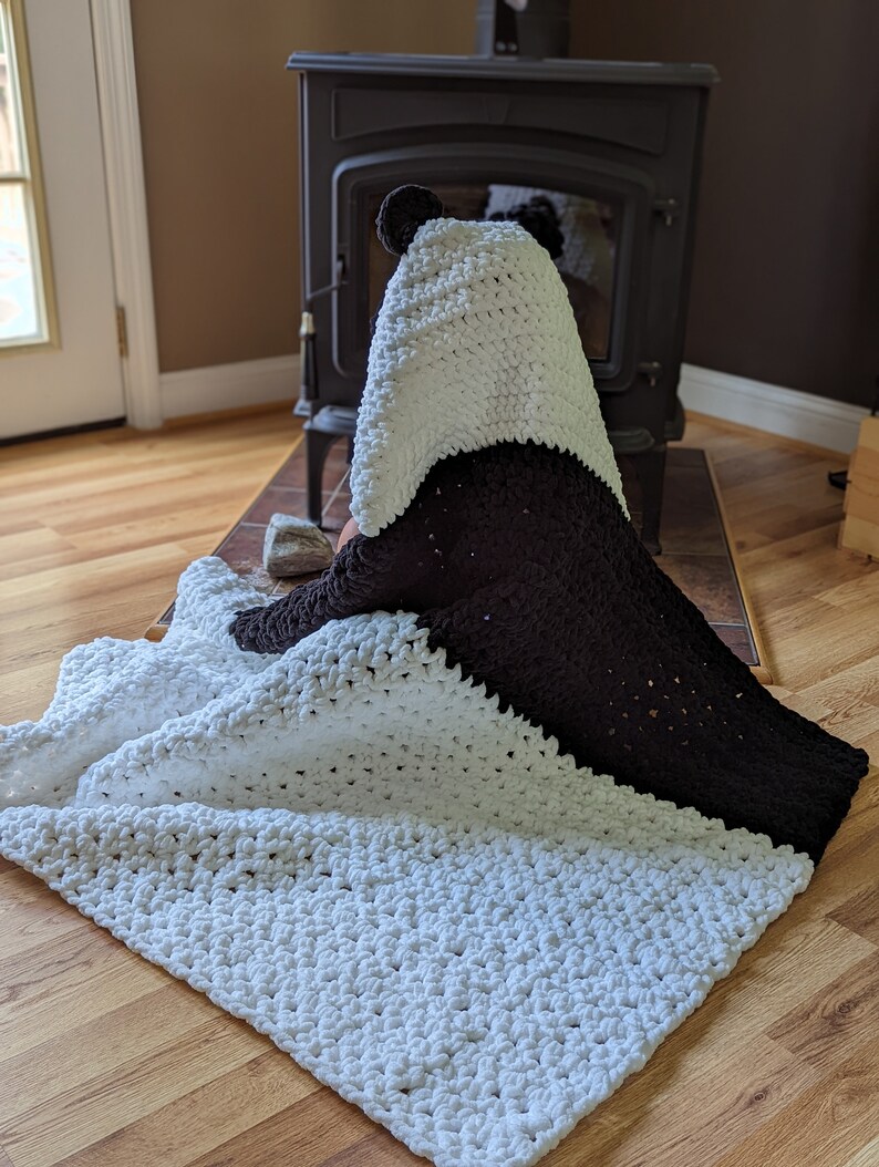 Puede incluir: Una manta con capucha de oso panda hecha a mano, blanca y marr&oacute;n. La manta est&aacute; hecha de un material suave y afelpado. La capucha presenta orejas de panda. La manta est&aacute; doblada sobre un suelo de madera.