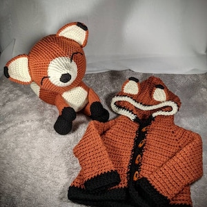 May include: A crocheted fox-themed set featuring a stuffed animal and a hooded jacket. The jacket is orange with black trim and buttons, and the hood has fox ears. The stuffed fox is orange, white, and black.