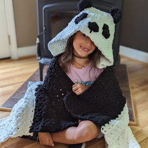 Puede incluir: Una manta con capucha de panda en blanco y negro. La capucha presenta orejas y detalles faciales de panda. La manta est&aacute; envuelta alrededor de una persona sentada en un suelo de madera. La manta est&aacute; hecha de un material suave y afelpado.