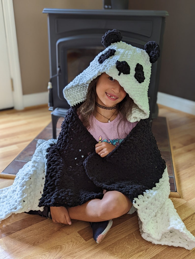 Puede incluir: Una manta con capucha de panda en blanco y negro. La capucha presenta orejas y detalles faciales de panda. La manta est&aacute; envuelta alrededor de una persona sentada en un suelo de madera. La manta est&aacute; hecha de un material suave y afelpado.