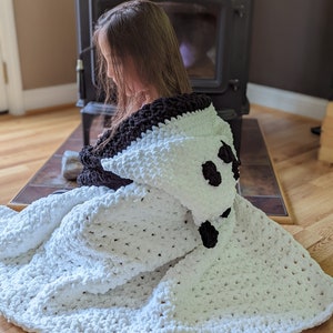 Puede incluir: Una manta con capucha blanca y negra con un dise&ntilde;o de panda. La manta est&aacute; hecha de un material suave y afelpado. La capucha presenta orejas y ojos negros. La manta est&aacute; sobre una persona.