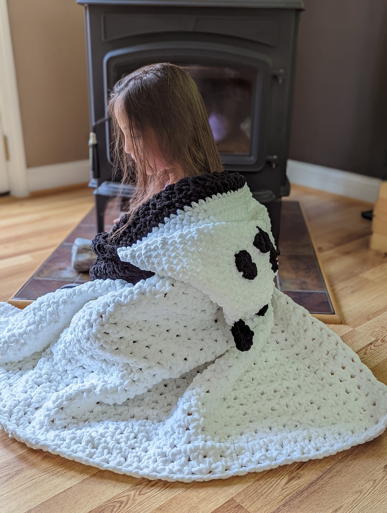 Puede incluir: Una manta con capucha blanca y negra con un dise&ntilde;o de panda. La manta est&aacute; hecha de un material suave y afelpado. La capucha presenta orejas y ojos negros. La manta est&aacute; sobre una persona.