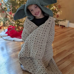 Puede incluir: Un ni&ntilde;o con un gorro con capucha verde con orejas grandes y envuelto en una manta beige de ganchillo. La manta es gruesa y texturizada. Un &aacute;rbol de Navidad decorado es visible en el fondo.