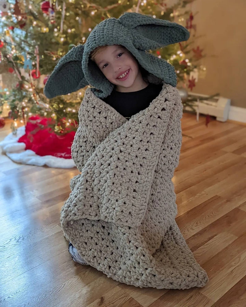 Puede incluir: Un ni&ntilde;o con un gorro con capucha verde con orejas grandes y envuelto en una manta beige de ganchillo. La manta es gruesa y texturizada. Un &aacute;rbol de Navidad decorado es visible en el fondo.
