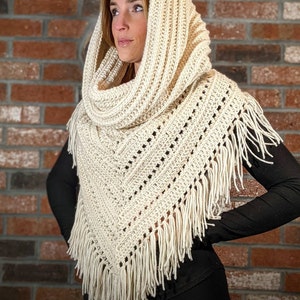 May include: Cream-colored crochet shawl with a hood and fringe. The shawl is made of thick yarn and features a geometric pattern with small holes. The fringe hangs along the bottom edge. The hood is draped over the head.