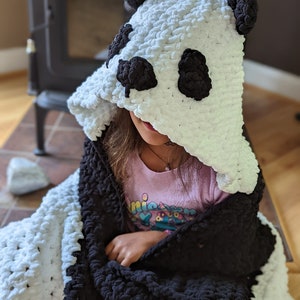 Puede incluir: Una manta con capucha hecha a mano con forma de oso panda. La manta es blanca con detalles negros, incluyendo la cara y las orejas del panda. El material suave y afelpado es visible. La manta est&aacute; dise&ntilde;ada para ni&ntilde;os.