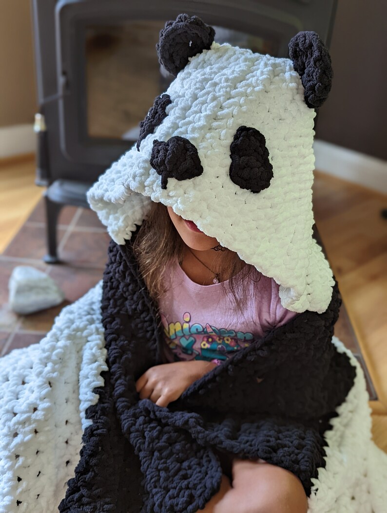 Puede incluir: Una manta con capucha hecha a mano con forma de oso panda. La manta es blanca con detalles negros, incluyendo la cara y las orejas del panda. El material suave y afelpado es visible. La manta est&aacute; dise&ntilde;ada para ni&ntilde;os.