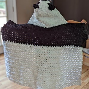 Puede incluir: Una manta con capucha de oso panda hecha a mano. La manta es blanca con una banda y capucha marr&oacute;n oscuro, con orejas y ojos negros. Hecha de un material suave y texturizado, ideal para la comodidad.