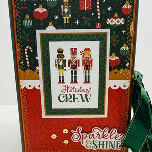 May include: A rectangular holiday-themed scrapbook album featuring a dark green background with nutcracker and ornament patterns, a central white panel with three nutcracker illustrations and the text Holiday CREW, and a red lower section with the text Sparkle & SHINE, finished with a green ribbon.