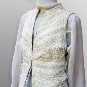 May include: A white and gold tweed waistcoat with a fringe trim. The waistcoat has a V-neck and a sleeveless design.