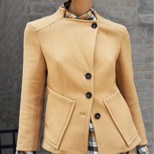 Könnte beinhalten: Hellbraune Jacke mit Stehkragen und drei dunklen Knöpfen. Die Jacke hat zwei Vordertaschen und eine taillierte Passform. Ein kariertes Hemd ist darunter sichtbar. Die Jacke wird auf einer Schaufensterpuppe präsentiert.