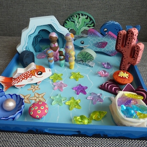 May include: A wooden mermaid-themed playset on a blue tray. Includes mermaids, fish, starfish, coral, and a pearl in a shell. The set features bright colors and whimsical designs, perfect for imaginative play.