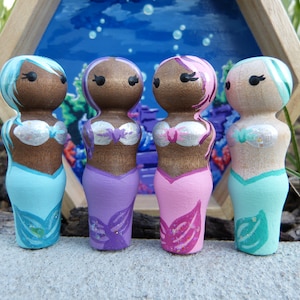 Puede incluir: Cuatro figuras de sirenas de madera con detalles pintados. Cada una tiene un cuerpo de madera marrón, un top de bikini plateado con purpurina y una cola colorida. Las sirenas tienen colas azules, moradas y rosas y pelo a juego. Se encuentran frente a una escena submarina azul.