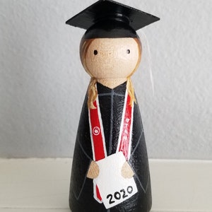 May include: Wooden peg doll dressed in a black graduation gown and cap. The doll has blonde hair and a red and white sash. It holds a white paper with the number "2020" on it. The doll is a graduation-themed item.