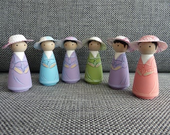 May include: Six wooden peg dolls, each painted in a different pastel color, including lavender, blue, green, and pink. The dolls have painted faces, dark hair, and wear dresses with decorative details. Each doll has a crocheted hat.