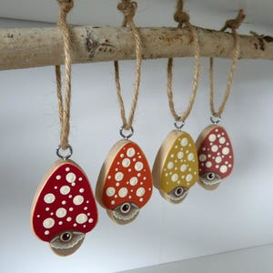 Puede incluir: Cuatro adornos de madera con forma de seta colgados de una rama natural con cuerda de yute. Cada seta presenta un sombrero pintado con puntos blancos y un ojo en la base. Los sombreros son de colores rojo, naranja y amarillo con herrajes.