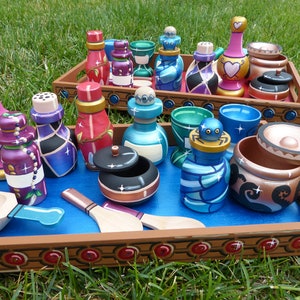 Puede incluir: Una colección de botellas, tarros y cuencos de madera pintados a mano en varias formas y tamaños, exhibidos en dos bandejas de madera. Los artículos presentan colores vibrantes, patrones decorativos y motivos de corazón. Algunos tienen tapas o tapones.