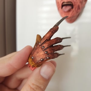 Puede incluir: Un conjunto de figuras esculpidas que presenta un rostro humano texturizado de color carne con ojos azules y la boca abierta con la lengua fuera, junto a un guante de color cobre metálico con cuatro dedos largos de hoja de metal afilados.