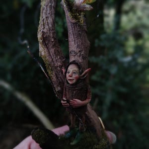 May include: A detailed miniature sculpture of a woodland sprite, crafted from clay, perched on a small tree branch. The figure is dressed in brown clothing and has a smiling face. The sculpture is set against a blurred green background.