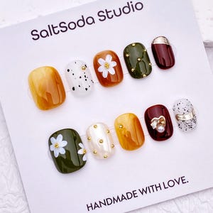 Op de afbeelding: Een set van tien kunstnagels op een witte SaltSoda Studio displaykaart. De nagels hebben diverse ontwerpen met amberkleurige, olijfgroene en donkerrode tinten, witte spikkels, kleine gouden studs, witte bloemaccenten en een parelbedel.