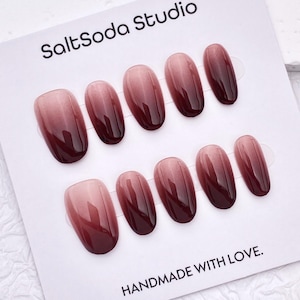 Peut inclure: Un ensemble de dix faux ongles disposés sur une carte de présentation blanche marquée SaltSoda Studio et HANDMADE WITH LOVE. Chaque ongle présente une finition brillante avec un dégradé ombré passant d'un vieux rose clair à un bordeaux profond.