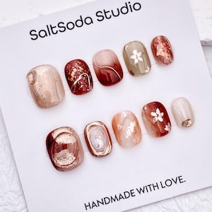 Op de afbeelding: Een set van tien kunstnagels op een witte displaykaart met het label SaltSoda Studio en HANDMADE WITH LOVE. De nagels hebben een mix van donkerrode, beige en saliegroene tinten met goudfolie accenten, metallic lijndetails en kleine bloemmotieven.