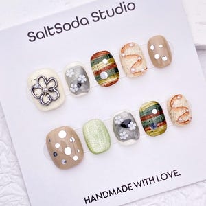 Op de afbeelding: Een set van tien press-on nagels op een witte displaykaart met het label SaltSoda Studio en HANDMADE WITH LOVE. De nagels hebben diverse ontwerpen, waaronder crème, groen en gestreepte patronen, geaccentueerd met zilveren bedels, bloemen en parels.