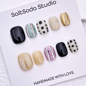 May include: A set of press-on nails in various designs, including black, striped, polka dot, and textured patterns. The nails are arranged on a white card with the text "SaltSoda Studio" and "HANDMADE WITH LOVE."