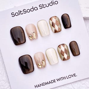 May include: A set of ten press-on nails on a white display card labelled SaltSoda Studio and HANDMADE WITH LOVE. The nails feature a mix of dark brown, cream, and nude colours with gold foil accents and a brown and cream argyle diamond pattern.