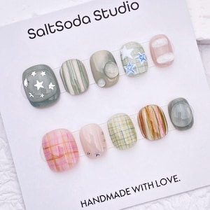 May include: A set of ten press-on nails on a white display card labeled SaltSoda Studio and HANDMADE WITH LOVE. The nails feature various designs including plaid patterns, star motifs, and solid colors in shades of sage green, pink, and tan.