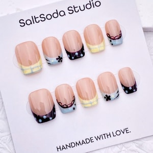 Può includere: Un set di unghie finte con base nude e vari disegni. Alcune unghie presentano un motivo a pois bianchi e neri, mentre altre hanno un accento floreale o un bordo giallo e blu. La carta dice "SaltSoda Studio" e "HANDMADE WITH LOVE."