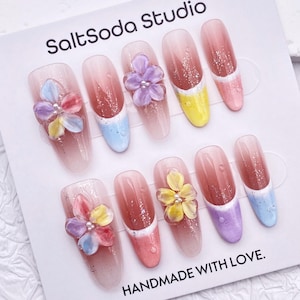 Peut inclure: Un ensemble de faux ongles avec un dégradé, ornés de fleurs en 3D. Les ongles présentent des couleurs variées, dont le rose, le jaune, le bleu et le violet, avec des paillettes. La carte indique "SaltSoda Studio" et "HANDMADE WITH LOVE."