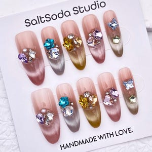 Peut inclure: Dix faux ongles avec une base rose clair et un dégradé. Chaque ongle est orné de strass et de pierres précieuses colorées. La carte indique "SaltSoda Studio" et "HANDMADE WITH LOVE."