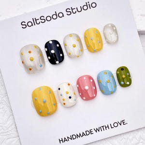 May include: A set of ten press-on nails displayed on a white card labelled SaltSoda Studio and HANDMADE WITH LOVE. The nails feature various glossy colours including yellow, black, white, pink, blue, and olive green, all decorated with small polka dots.