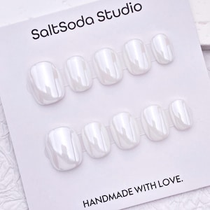 Könnte beinhalten: Ein Set aus zehn künstlichen Fingernägeln mit glänzendem, perlmuttweißem Finish, angeordnet in zwei Reihen auf einer weißen Präsentationskarte. Die Karte zeigt oben den Text SaltSoda Studio und unten HANDMADE WITH LOVE. in schwarzer Schrift.