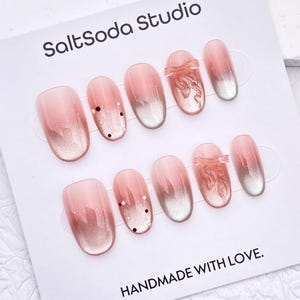 Peut inclure: Un ensemble de faux ongles avec un dégradé rose et argenté. Certains ongles présentent des petits points noirs ou un nœud décoratif. La carte indique "SaltSoda Studio" et "HANDMADE WITH LOVE."
