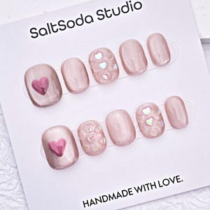 Könnte beinhalten: Ein Set aus hellrosa Kunstnägeln mit Herz- und Glitzerdetails. Die Nägel sind auf einer weißen Karte mit dem Text "SaltSoda Studio" und "HANDMADE WITH LOVE." angeordnet.