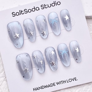 May include: A set of ten oval-shaped, light blue press-on nails. Each nail features silver star embellishments and a gradient effect. Some nails have a light blue bow design. The card reads "SaltSoda Studio" and "HANDMADE WITH LOVE."