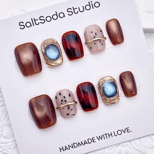 May include: A set of press-on nails with various designs. Some nails are a metallic brown, others have a tortoiseshell pattern, and some are a light pink with black speckles. Two nails feature a gold-colored frame with a blue stone. The card reads "SaltSoda Studio" and "HANDMADE WITH LOVE."