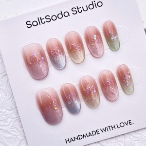 Peut inclure: Un ensemble de faux ongles ovales avec un dégradé de rose à argenté, puis beige clair. Les ongles sont ornés de paillettes irisées et présentés sur une carte blanche avec le texte "SaltSoda Studio" et "HANDMADE WITH LOVE."