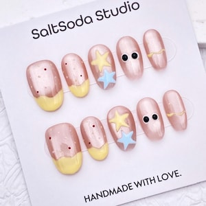 Peut inclure: Un ensemble de faux ongles avec une base rose nacrée. Certains ongles présentent des pointes jaunes, des points rouges ou des motifs d'étoiles et d'yeux. La carte indique "SaltSoda Studio" et "HANDMADE WITH LOVE."