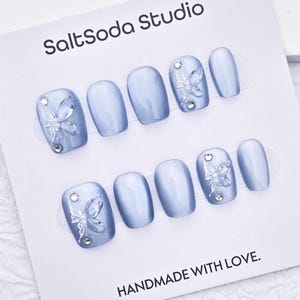 May include: A set of light blue press-on nails with butterfly and rhinestone accents. The nails are displayed on a white card with the text "SaltSoda Studio" and "HANDMADE WITH LOVE."