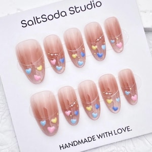 Peut inclure: Dix faux ongles avec un dégradé nude et marron. Chaque ongle est orné de petits cœurs pastel roses, jaunes et bleus, ainsi que d'une délicate décoration perlée. La carte indique "SaltSoda Studio" et "HANDMADE WITH LOVE."
