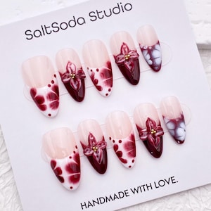Pode incluir: Um conjunto de unhas postiças com base rosa claro, um design marmorizado em vermelho escuro e branco e um detalhe floral em vermelho escuro com detalhes dourados. O cartão diz "SaltSoda Studio" e "HANDMADE WITH LOVE."