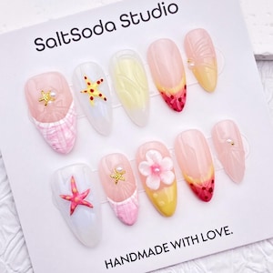 Peut inclure: Un ensemble de dix faux ongles avec divers motifs. Certains ongles présentent du tartan rose et blanc, des étoiles de mer, des fleurs et des accents dorés. Les ongles sont présentés sur une carte blanche avec le texte "SaltSoda Studio" et "HANDMADE WITH LOVE."