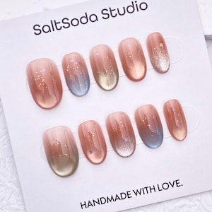 Peut inclure: Un ensemble de faux ongles ovales avec un dégradé de couleur, passant du marron clair au bleu et paillettes argentées. La carte porte l'inscription "SaltSoda Studio" et "HANDMADE WITH LOVE."