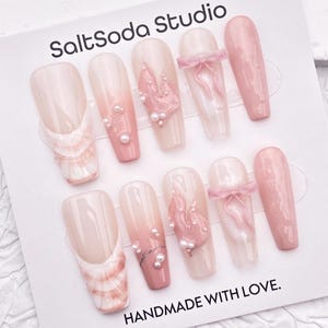May include: A set of ten artificial nails in various designs. The nails feature a range of colors, including pink, white, and nude. Some nails have pearl accents, bows, and textured designs. The card reads "SaltSoda Studio" and "HANDMADE WITH LOVE."