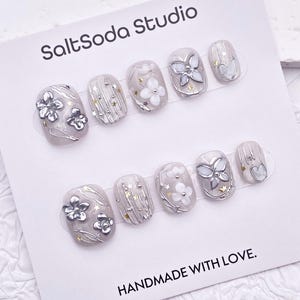 以下が含まれることがあります： SaltSoda Studioの楕円形のつけ爪セット。爪は明るいグレーを基調とし、銀色の花柄と蝶のデザイン、金色の星のアクセントが特徴です。「HANDMADE WITH LOVE」と書かれたカード。